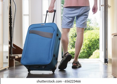 Tourist Young Man In T-shirt, Shorts And Sandals With Blue Vacation Bag Exits Through The Front Door.