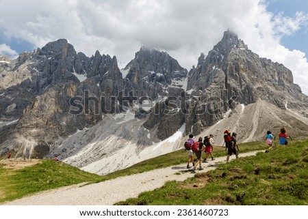 Tourists hiking on a track towards Cimon della Pala amid rugged peaks of Pale di San Martino mountain range in Dolomites on a sunny summer day, in Passo Rolle, Trentino Alto Adige, South Tyrol, Italy