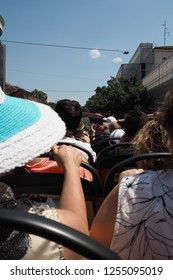 Tourists Seated On The Open Top Deck Of A Tour Bus In Rome, Italy, With Buildings And Blue Sky