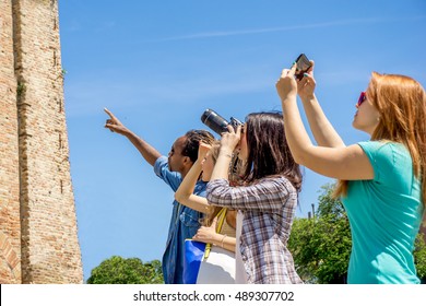 tourists sightseeing taking photos with reflex camera and smart-phones in front of a monument. group of multi-ethnic foreign visitors looking at a historic building - man points finger and explains