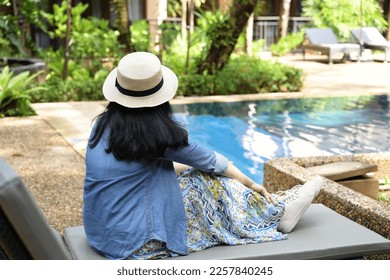 Tourists sit by the pool happily on vacation. A middle-aged woman with long hair wearing a blue flared skirt. In a wide-brimmed hat and a denim shirt. Sitting on a beach chair by the - Powered by Shutterstock