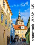 Tourists walking on a cobblestone street between colorful buildings under a blue sky are approaching the famous clock tower in Sighisoara, Romania, on a sunny winter day