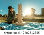Tower game. Happy family with kid playing board games outdoors in the park. Smiling mom and teen girl sitting on the floor and pulling wooden blocks from the tower