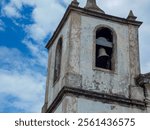 Tower of an old church, with worn paiting in a checkerboard shape and a rusty bell hanging witn blue sky and clouds