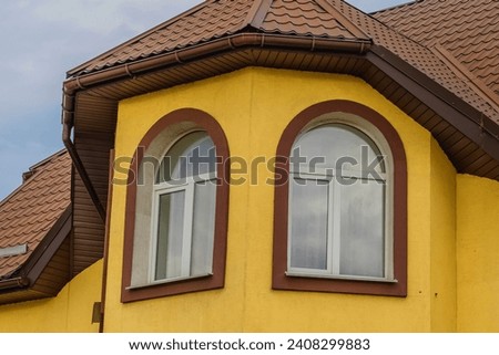 It is  tower with windows on a top floor of European house with bright yellow walls and brown tilled roof.  There is achimney on roof. It is cloudy day It is close up view pf building