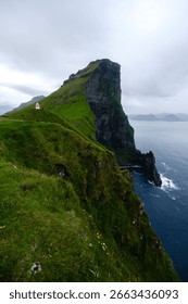 Towering cliffs rise dramatically from the Atlantic Ocean in the Faroe Islands, enveloped in lush green grass. The overcast sky adds a mystical charm to this Kallur Lighthouse, Kalsoy Island