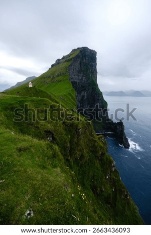 Towering cliffs rise dramatically from the Atlantic Ocean in the Faroe Islands, enveloped in lush green grass. The overcast sky adds a mystical charm to this Kallur Lighthouse, Kalsoy Island