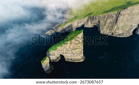 Towering Drangarnir cliffs rise dramatically from the dark waters of the Atlantic, adorned with vibrant green foliage. Mist envelops the rocks, creating a mystical atmosphere in the Faroe Islands.