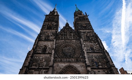 A towering Gothic cathedral with intricate stonework and twin spires reaching into a vibrant blue sky streaked with clouds. The grand facade is detailed with ornate carvings and a large rose window. - Powered by Shutterstock