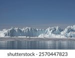 A towering ice wall of the Eqip Sermia Glacier in Greenland reflects in the calm waters of the fjord under a clear blue sky, showcasing jagged ice formations and glacial textures