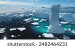 Towering melting iceberg in Arctic icy ocean. Snow covered glacier drift under blue sunny sky. Pure ice floats in turquoise water. Ecology, melting ice, climate change global warming. Aerial view