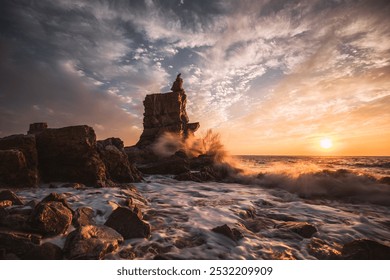 Towering rock formation meeting crashing ocean waves, set against a vibrant sunset sky with swirling clouds and warm sunlight reflections. - Powered by Shutterstock