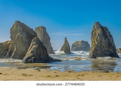 Towering sea stack columns loom over a wet reflective beach at low tide in Bandon, Oregon, with crashing surf and a vivid blue sky backdrop.