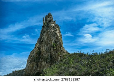 A towering, sharp rock pinnacle stands majestically on a grassy hill, reaching toward a dramatic blue sky filled with wispy white clouds, showcasing natural geological erosion and strength.
