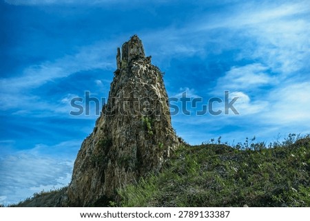 A towering, sharp rock pinnacle stands majestically on a grassy hill, reaching toward a dramatic blue sky filled with wispy white clouds, showcasing natural geological erosion and strength.