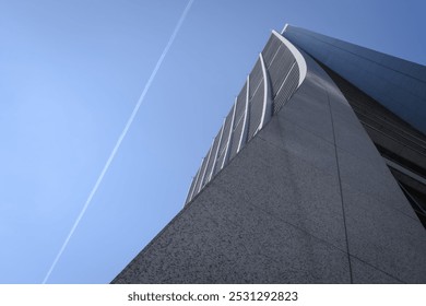 A towering skyscraper rises dramatically against a clear blue sky, showcasing its sleek, modern architecture. A contrail cuts across the sky, adding a dynamic element to the scene. - Powered by Shutterstock