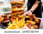 A towering triple-decker burger with crispy chicken patties, fresh lettuce, onions, and sauce, accompanied by golden fries and fried chicken wings on a black serving tray.