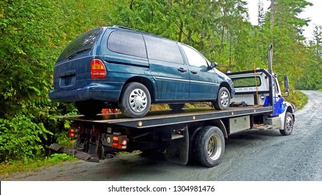 Towing Service - The Blue Tow Truck With The Loaded Old Damaged Car Which Stopped Working In The Middle Of Forest On The Rough Off Road. Back View