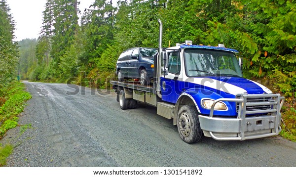 Towing service - the blue truck with the\
loaded old damaged mini van car which stopped working in the middle\
the forest on the rough off road. Front\
view