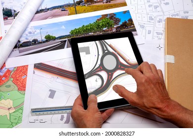 Town planning - Land use planning - Employee holding in his hands a digital tablet displaying a road project plan, above a desk where ground plan maps are placed 