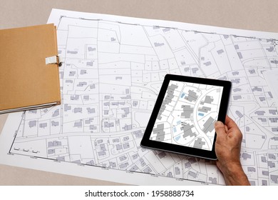 Town Planning And Land Use Planning - Hand Holding A Digital Tablet Displaying A Local Urban Plan, Above A Cadastral Plan Placed On A Desk