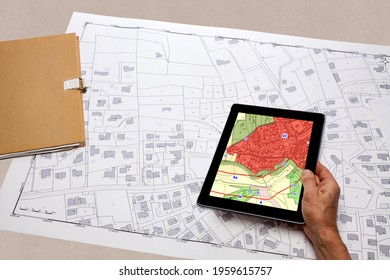 Town Planning And Land Use Planning - Hand Holding A Digital Tablet Displaying A Local Urban Plan, Above A Cadastral Plan Placed On A Desk