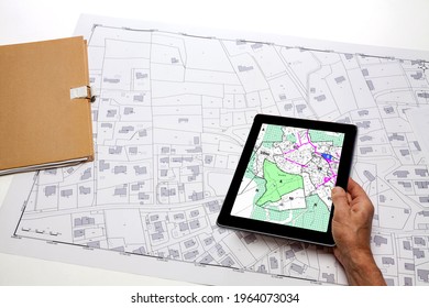 Town Planning And Land Use Planning - Hand Holding A Digital Tablet Displaying A Local Urban Plan, Above A Cadastral Plan Placed On A Desk