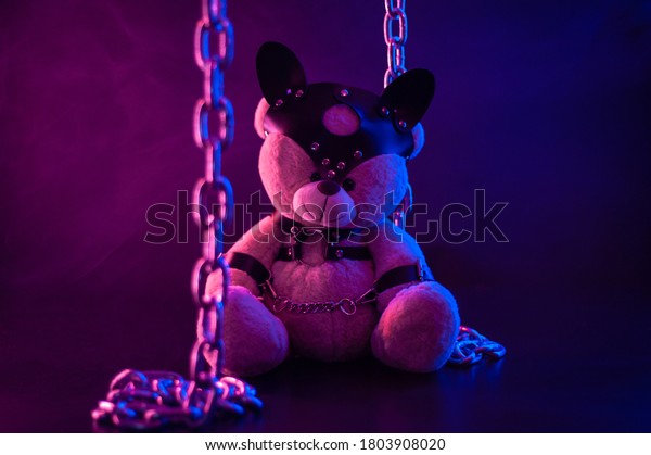 toy bear dressed in leather\
belts harness accessory for BDSM games on a dark background in neon\
light