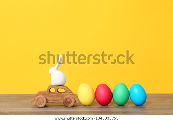 Toy car with ceramic bunny and Easter\
eggs on wooden table against color\
background