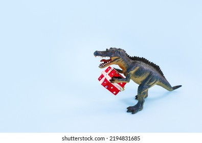 Toy Dino With A Plastic Gift Box On Blue Background. The Concept Of Congratulations On The Holidays. Selective Focus, Copy Space