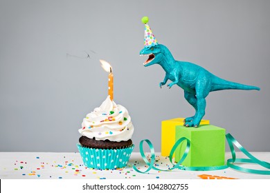 Toy Dinosaur blowing out a Birthday candle with cup cake on a gray background