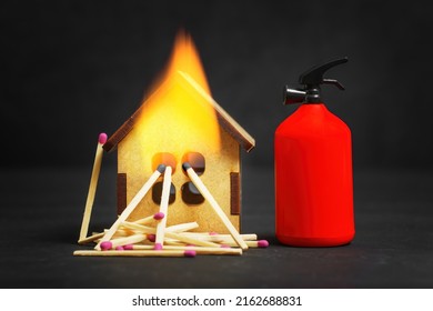 Toy Fire Extinguisher Placed By A Burning Tiny Wooden House Model On Black Background. Household Fire Safety Plan Concept.