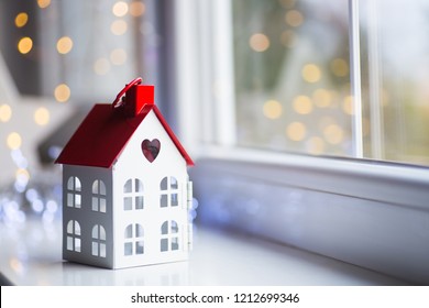 Toy House With Hole In Form Of Heart Near Window In Daylight With Garland Lights On Background. Romantic Card. Valentine Day Banner.