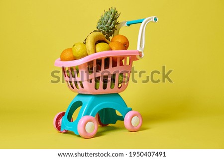 Toy kids pink shopping cart full of assorted exotic fruits isolated on yellow background with copy space. Summer concepts