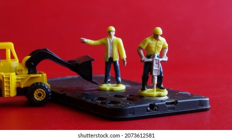 Toy Man Workers Recycle Or Repair An Old Mobile Phone Or Smartphone Using A Jackhammer And A Forklift Truck. Smartphone Recycling Or Repair. Red Background. Micro World. Selective Focusing. Close-up