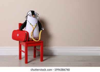Toy penguin with eyeglasses, stethoscope and first aid bag near beige wall, space for text. Pediatrician practice