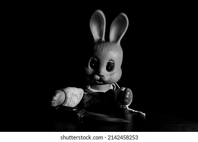 A Toy Rubber Bunny Sits With A Knife At The Table. Creepy Rabbit With Glowing Eyes