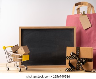 Toy Shopping Cart With Boxes With Mock Up Chalk Blackboard Frame On The Wooden Table With Bag And Gift Box. Gray Walls. Shopping Concept.