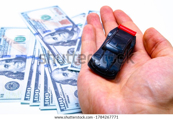 A toy\
sports car of black color with a red wing on in a man\'s hand on the\
background dollars. Car and dollars. Buying, selling price\
increases in the market July 2020. Los\
Angeles.USA