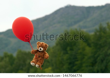 A toy teddy bear flies through the sky on a red ball, against a background of forest and mountains.                                