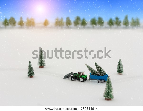 Toy tractor with a trailer carries Christmas trees\
during snowfall, rides through the snow in the middle of the\
forest. Beautiful background for greeting card. Winter composition.\
Happy holiday mood. 