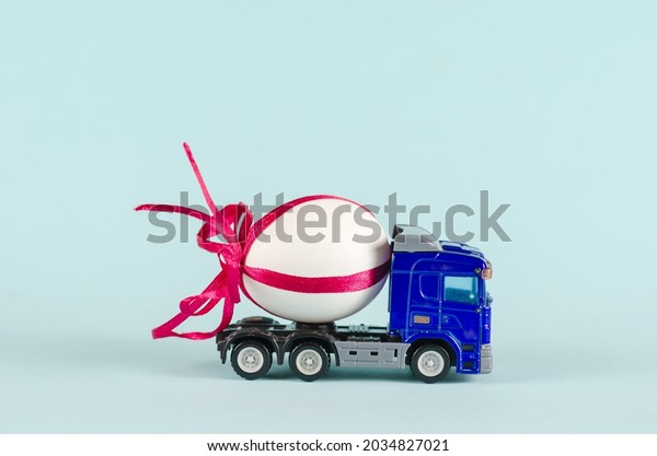 Toy tractor transports a chicken egg. Blue plastic\
truck and white egg with red ribbon. Blue background. Trucking.\
Selective focus.