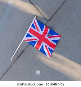 Toy Union Jack British Flag In Red, White And Blue Paper Fabric And White Stick Left Discarded On The Side Walk (pavement). The Cracks And Lines On The Concrete Create Diagonal Patterns And Shadows.