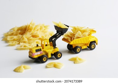 Toy Yellow Forklift Truck With A Bucket Loads Farfalle Paste Into The Back Of A Heavy-duty Dump Truck. Concept Of Delivering Food Products, Preparing For Sale Or Cooking. White Background. Daylight