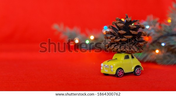 Toy yellow small car with pine cone on red\
background with blurred bokeh and Christmas tree branch. Winter\
holiday xmas theme, festive bright background. Close-up, selective\
focus with copy space area