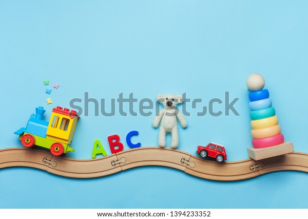 Toys background with\
copy space. Kids toys train, ABC letters, bear, car and pyramid on\
toy wooden railway on blue background with blank space for text.\
Top view, flat lay.