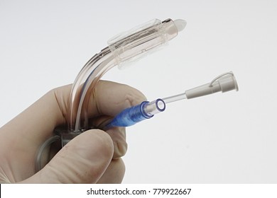 Tracheostomy Cannula With White Obturator Inserted And Channels For Inflanting And Suction Visible, Held In Left Hand In Powdered White Sterile Latex Glove, Light Grey Background