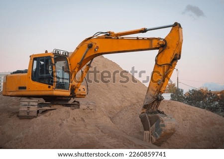 A tracked orange backhoe loader is parked on a large mountain of sand. A crawler excavator loads crushed stone and sand into a truck. Large hills and pillars of sand.
