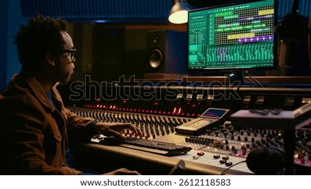 Tracking engineer processing and mixing sounds in control room with audio console and professional editing software on computer. Music production expert uses motorized faders and knobs. Camera B.