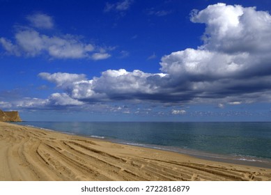 “Tire Tracks on Sandy Beach with Dramatic Clouds”

“A tranquil coastal scene featuring a sandy beach marked by tire tracks, leading the viewer’s eye toward a calm ocean horizon. On the left, a rocky cliff juts into the water, adding geological contrast to the soft textures of sand and sea. Above, dramatic cumulus clouds dominate the sky, casting shadows across the beach and ocean while allowing glimpses of blue. The interplay of textures — from the granular sand to the smooth water and layered clouds — creates a visually compelling and serene composition, ideal for themes of travel, solitude, and natural beauty.”